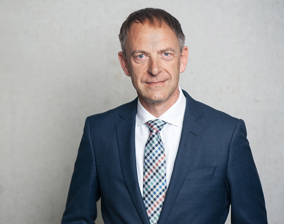 Christian Buck im dunkelblauen Anzug mit karierter Krawatte vor einer grauen Betonwand