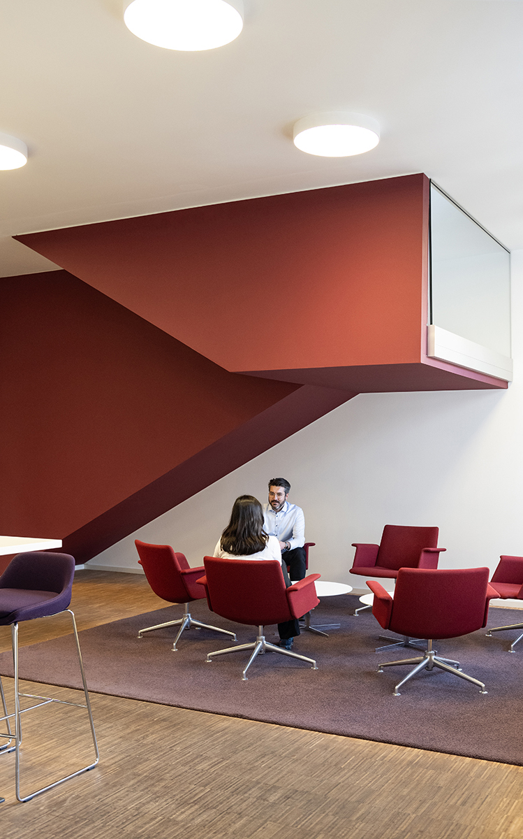 Zwei Personen im Gespräch auf roten Sesseln unter einer roten Treppenskulptur