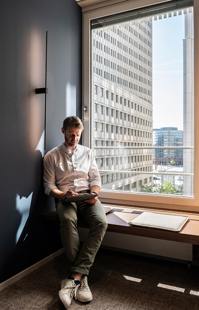 Mann mit Tablet auf einer Fensterbank vor Hochhausfassade