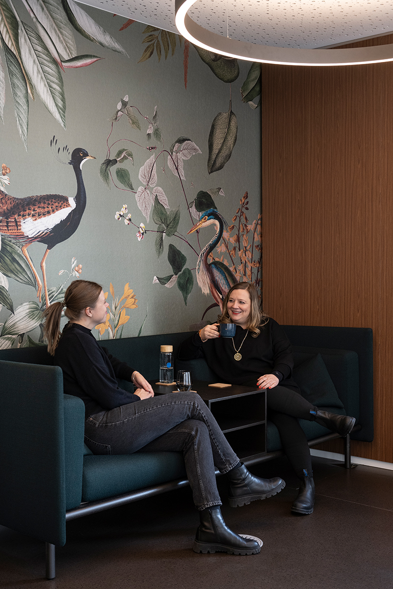 Zwei Frauen beim Kaffee auf dunkelgrünen Sofas vor einer Tapete mit Vögeln und Pflanzen