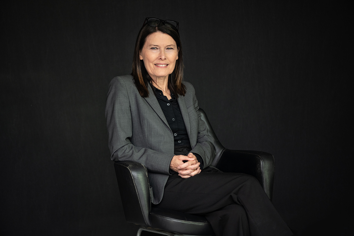 Cordula Heckmann im grauen Blazer, lachend auf einem Ledersessel vor schwarzem Hintergrund
