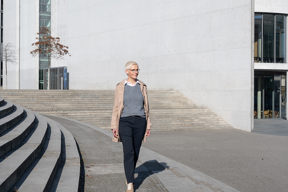 Kati Fink im Trenchcoat, gehend vor einer hellen Betonfassade mit geschwungenen Stufen