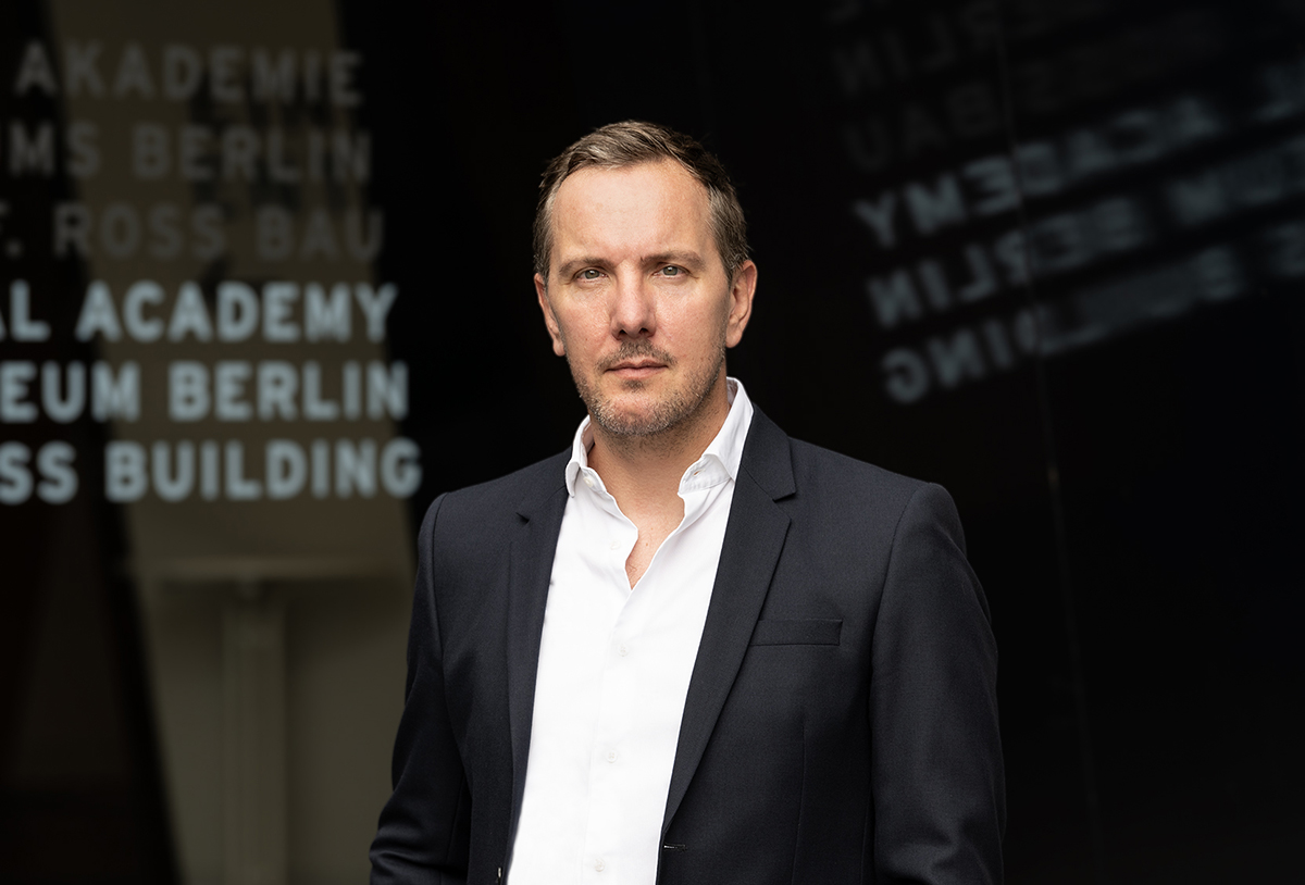 Thomas Helbing im dunklen Sakko und weißen Hemd vor einer dunklen Glasfläche mit Schriftzügen