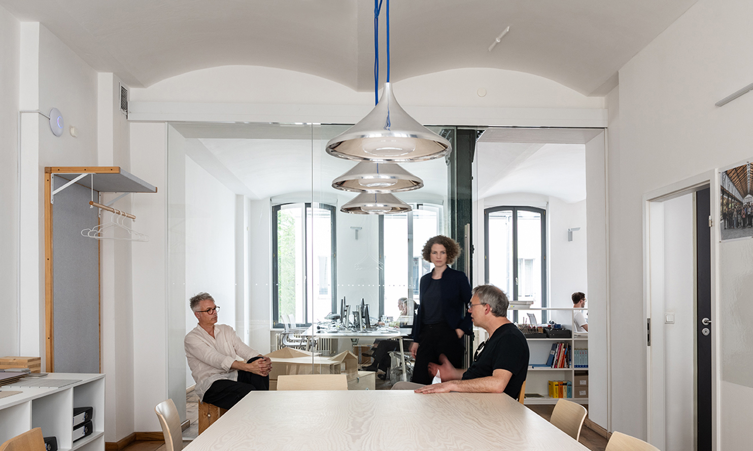 Drei Personen im Besprechungsraum des Architekturbüros unter drei silbernen Pendelleuchten, im Hintergrund das Büro